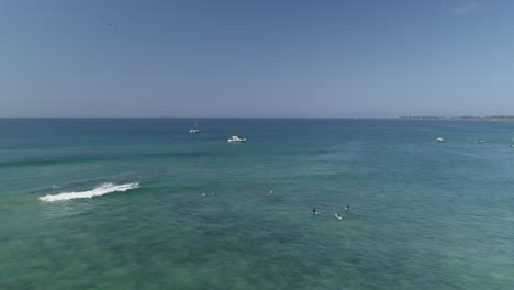 Aerial-orbiting-surfers-in-blue-water-with-small-boats-in-background,-4K