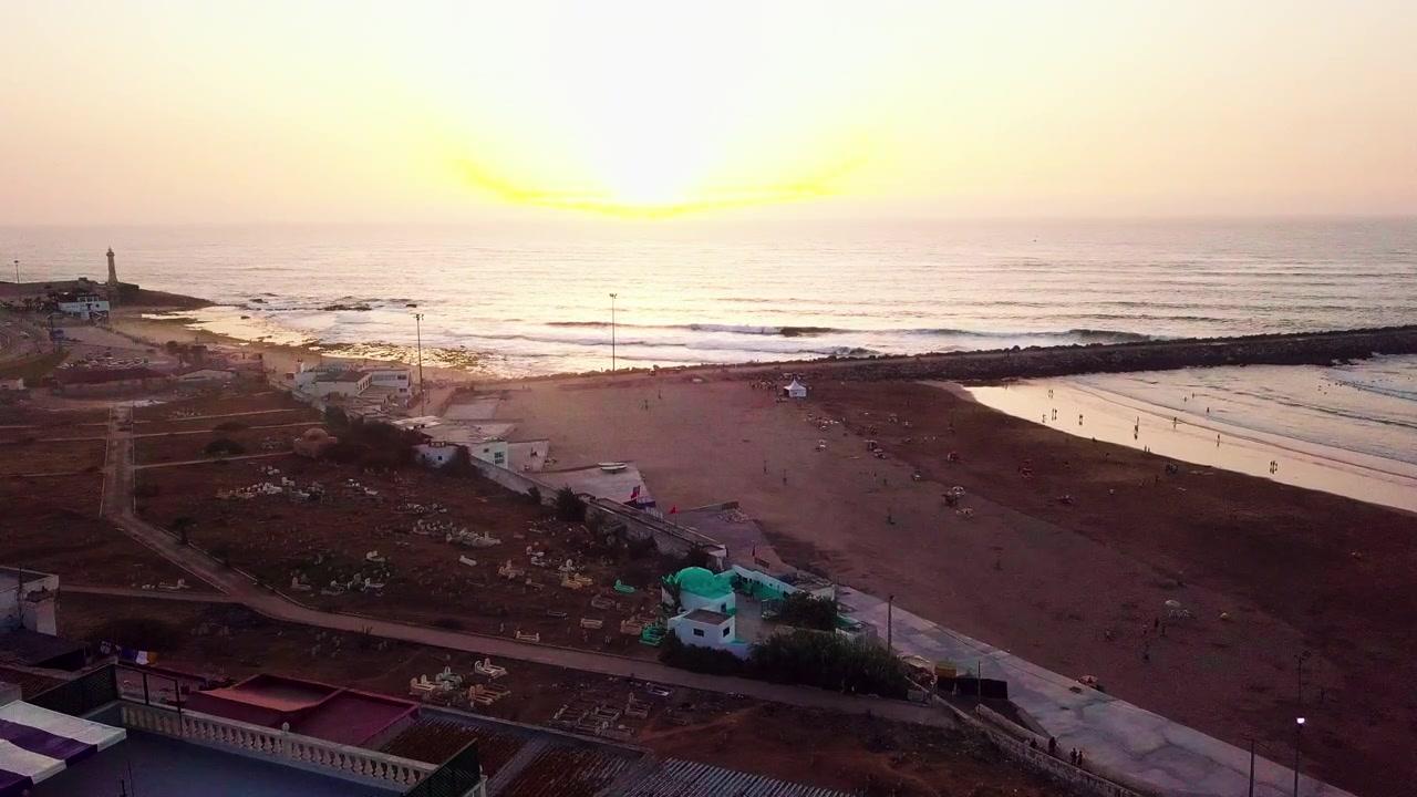 Dron sobrevolando los tejados junto al mar hacia una playa de arena en Marruecos al atardecer, con olas tranquilas, una cálida luz dorada y un ambiente costero apacible