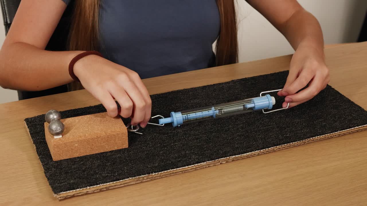 A person conducts a friction experiment using a spring scale, weights, and a textured surface on a table