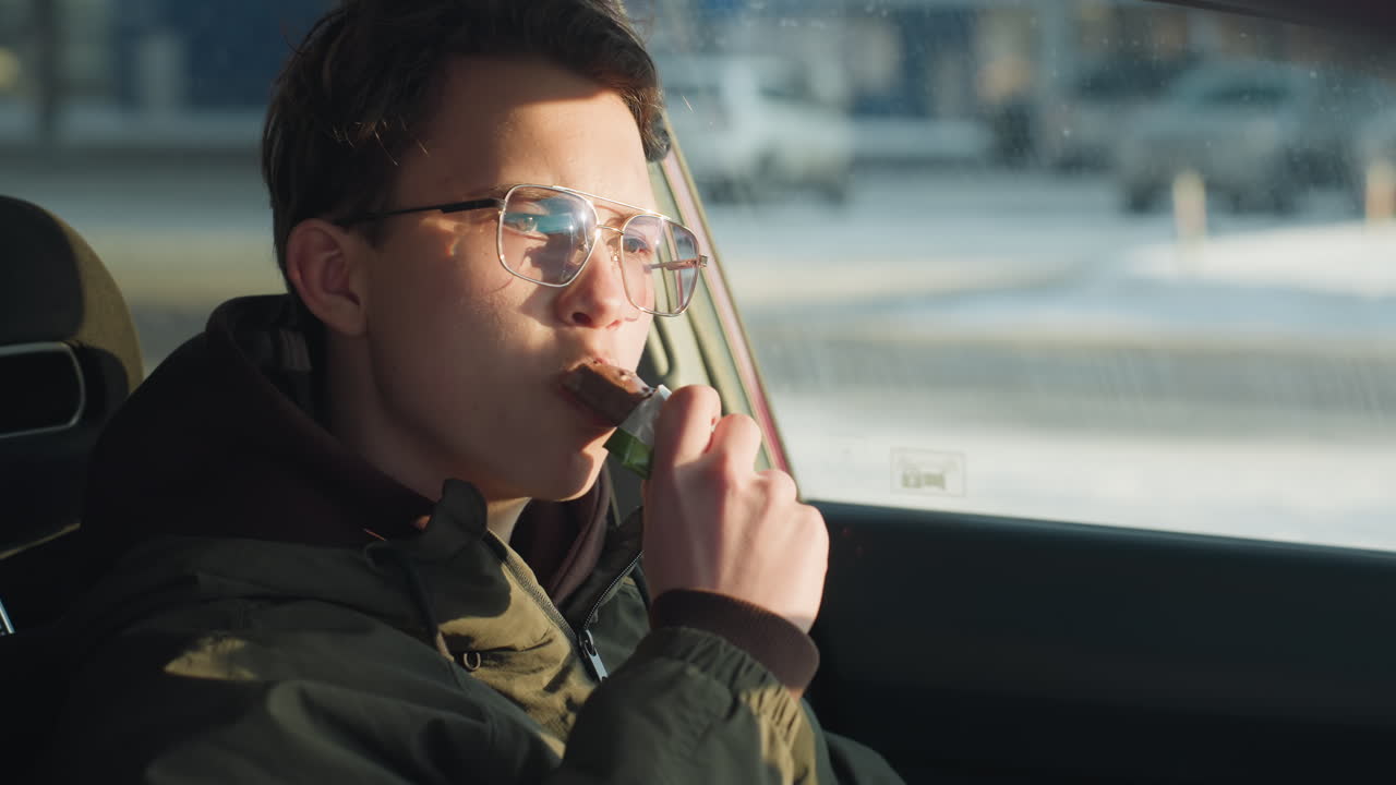 close up of young boy savoring chocolate candy inside parked car with sunlight reflecting off his face and industrial building blurred in background conveying warm sweet indulgence in winter