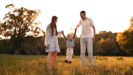 Mamá-Y-Papá-Sacuden-Al-Niño-Que-Juega-Con-Sus-Manos,-El-Niño-Está-Feliz-Jugando-Con-Sus-Padres-En-La-Calle-En-El-Verano-Al-Atardecer.