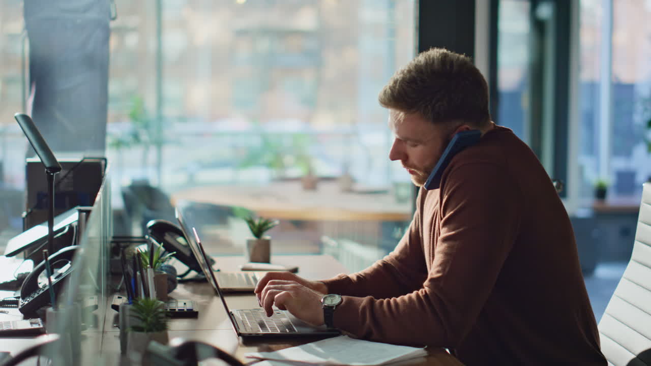 Überbeschäftigter Angestellter spricht am Handy, während er auf dem Arbeitsplatz einen Laptop tippt