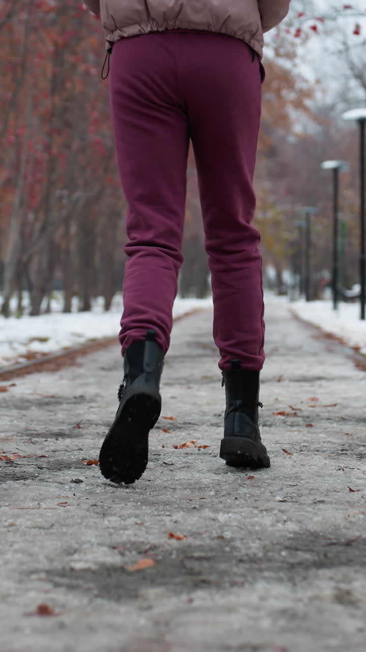 primer plano de las piernas y botas de una mujer joven corriendo por un camino cubierto de nieve helada en traje de invierno, en un parque al aire libre con árboles alineados y bayas rojas