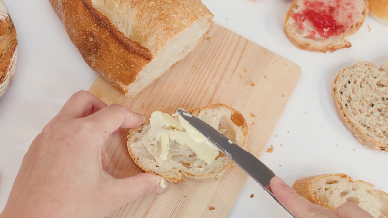Una persona spalma burro cremoso su pane di pasta madre croccante usando un coltello su un tagliere di legno.