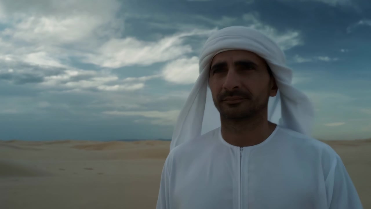 Vista frontal amplia de um retrato do peito para cima para mostrar mais do deserto ao redor enquanto o homem permanece centralizado vestindo roupas brancas, com luz do dia suave, dunas baixas e nuvens em camadas no horizonte