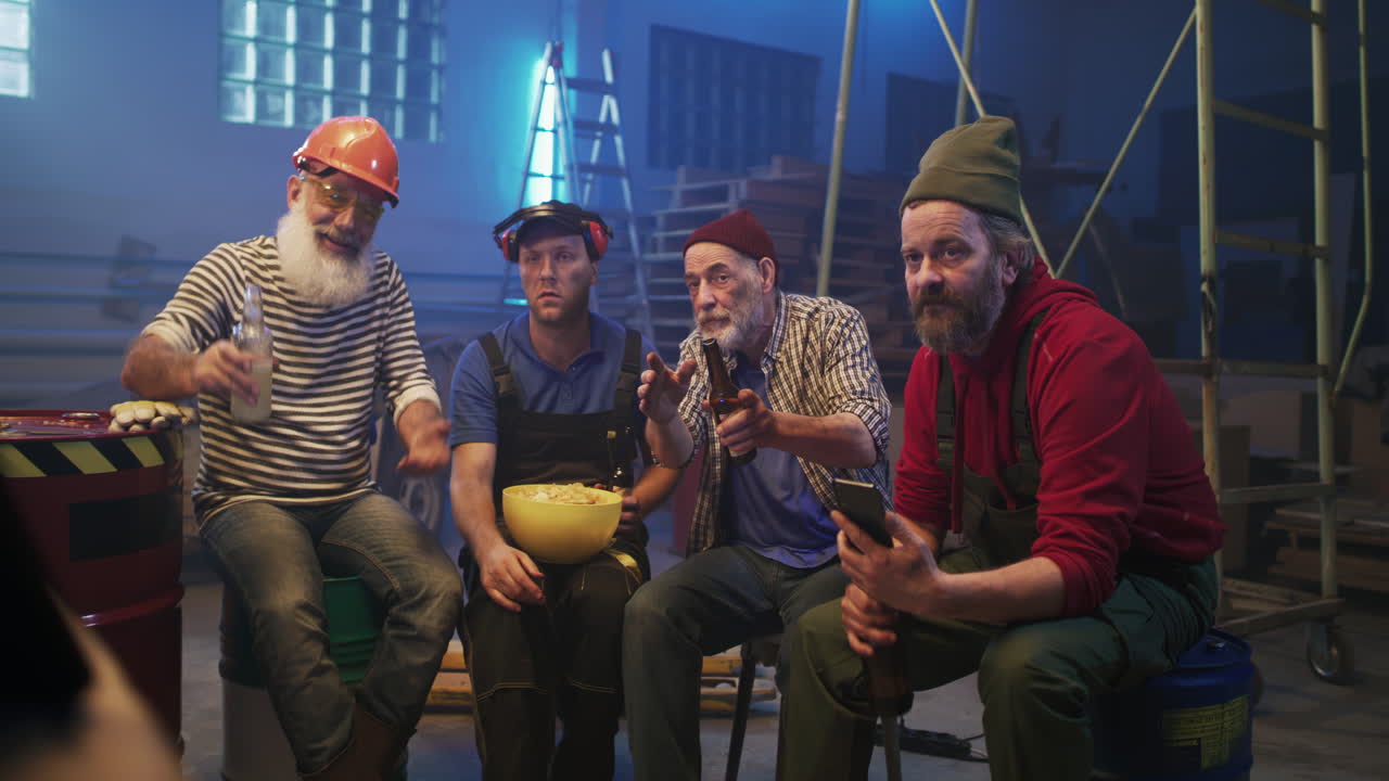 A group of mature men socialize, drink beer, and watch a game on a phone in a dimly lit workshop