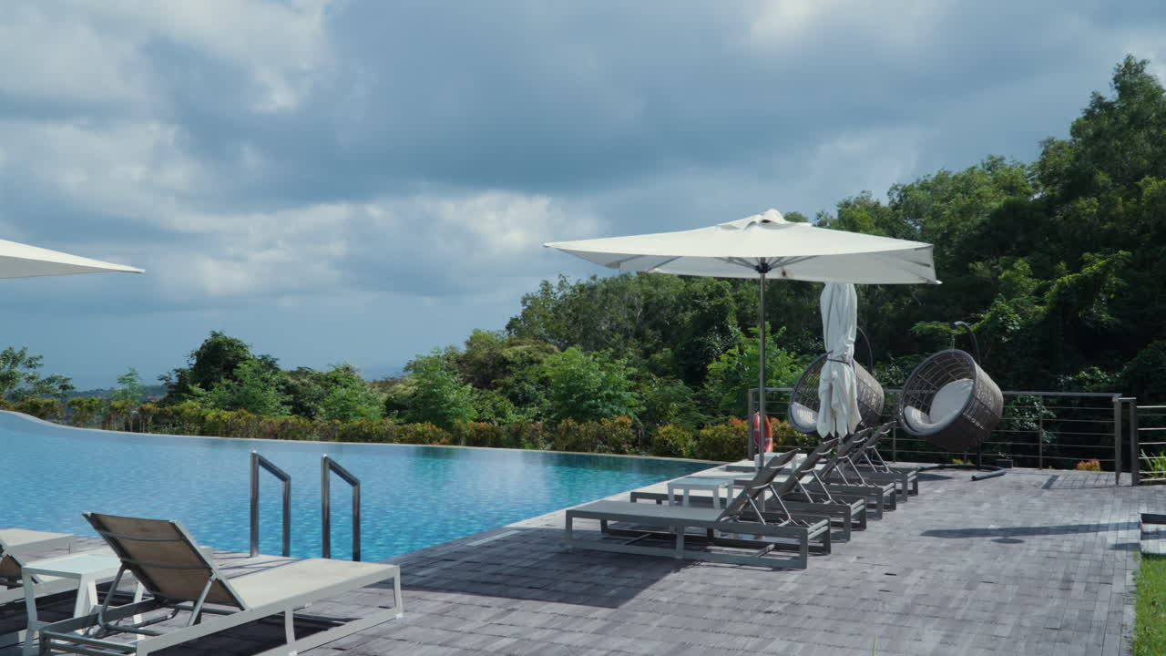 Sunbeds With Umbrellas On The Poolside Of Resort Villas During Cloudy Day In Bali, Indonesia. Wide Shot