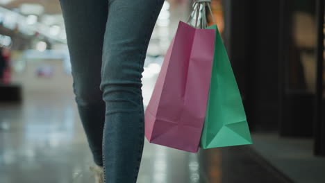 close-up leg view of a woman in jeans and canvas sneakers carrying mint and pink shopping bags in her right hand while walking through a brightly lit shopping mall with glossy tiled floors