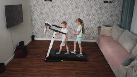 boy glances back while walking on treadmill with focused girl behind him in cozy living room setting with patterned wall and wooden flooring, both kids dressed in activewear during home workout