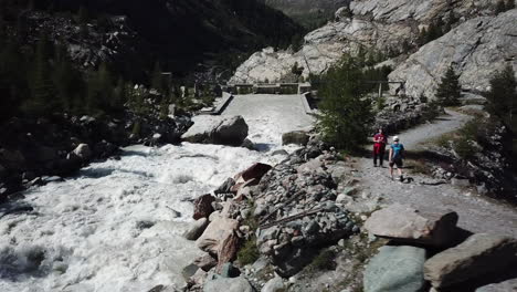 vista aérea de drones de un torrente fangoso en los alpes suizos junto a un bosque de abetos y un camino de grava