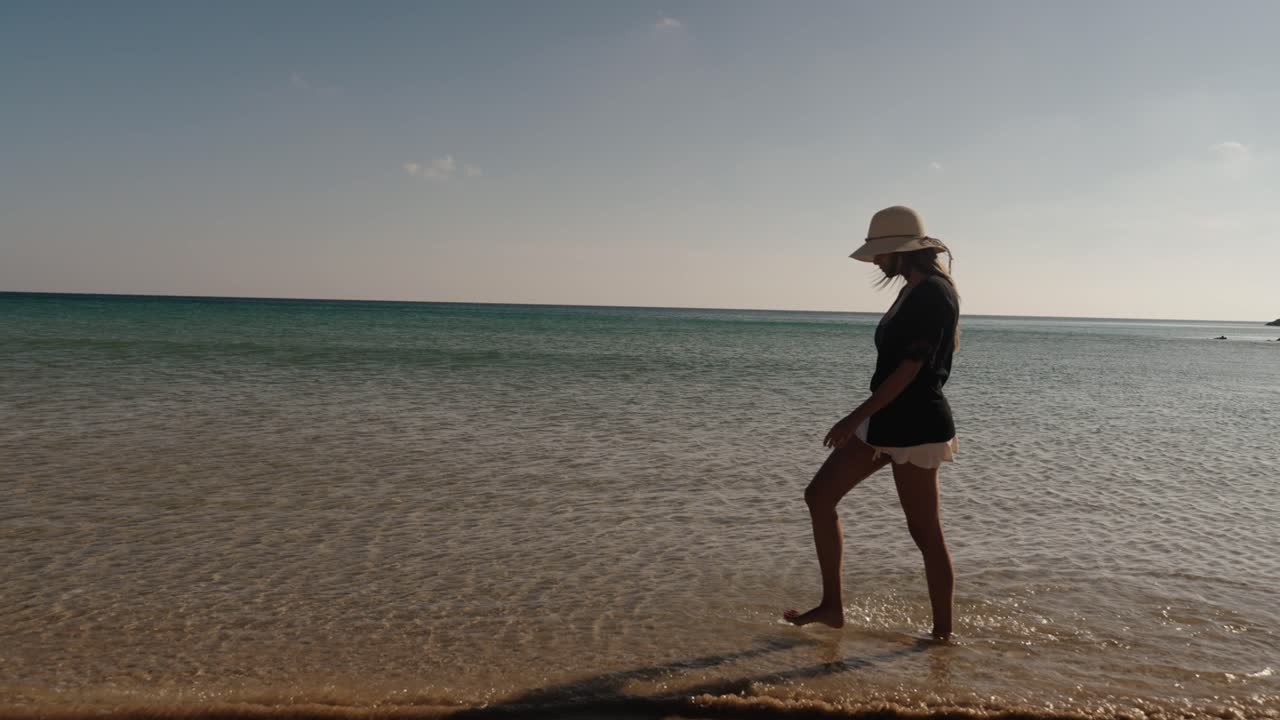 A woman strolls barefoot through shallow waves on a sunny beach in Algarve, Portugal, wearing a summer hat