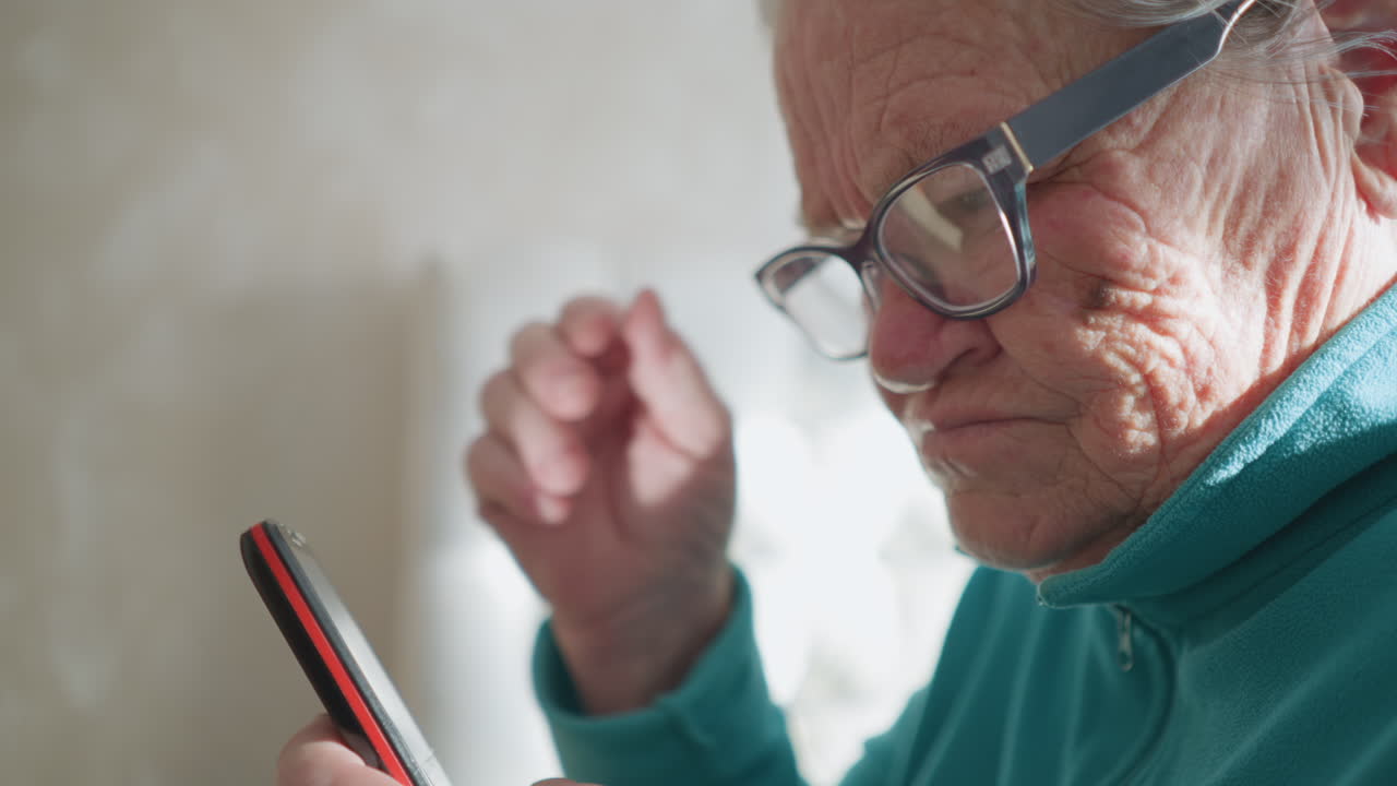 Elderly woman wearing turquoise fleece sweater, holding smartphone in one hand, adjusting glasses with the other. Focused expression, sunlight casting soft glow in background