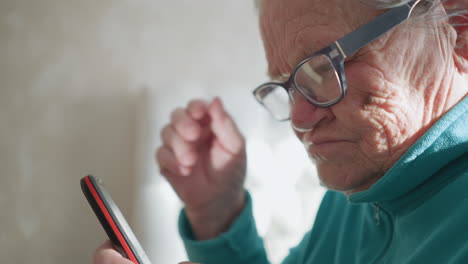 elderly woman wearing turquoise fleece sweater, holding smartphone in one hand, adjusting glasses with the other. focused expression, sunlight casting soft glow in background