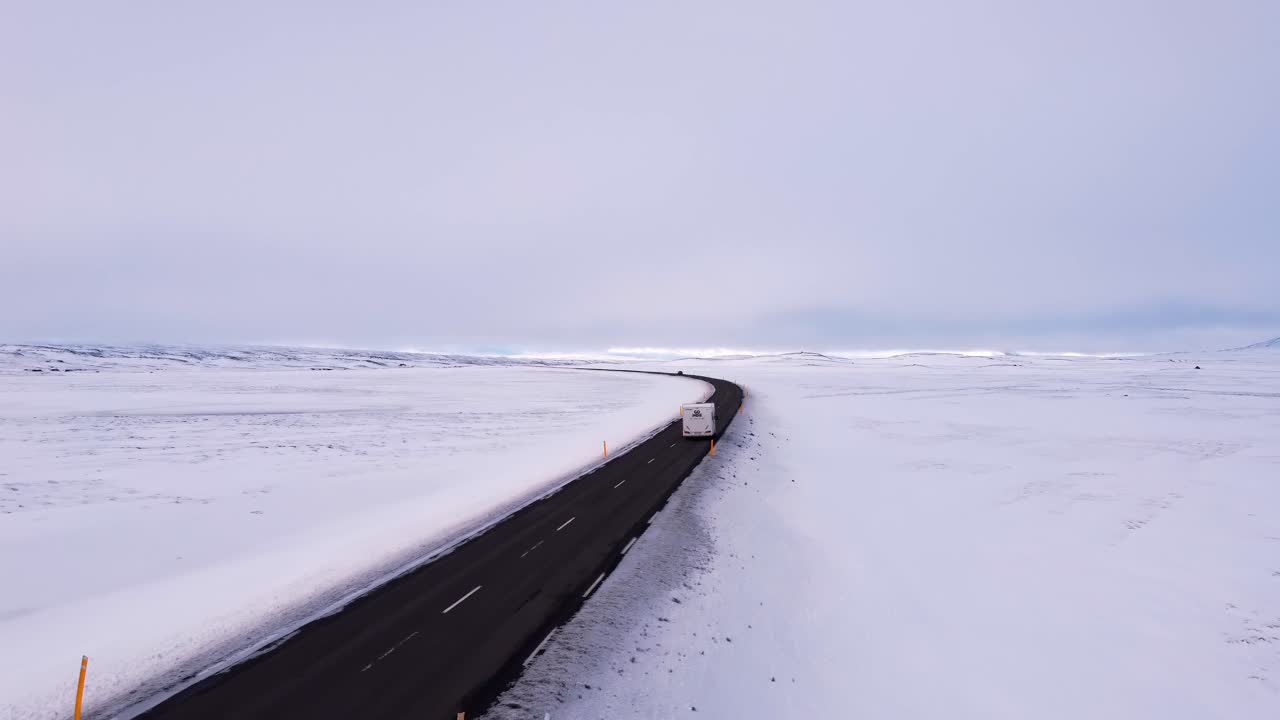A wide aerial shot captures a motorhome traveling along a dark asphalt road winding through an expansive, desolate winter landscape