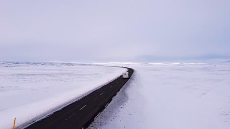 a wide aerial shot captures a motorhome traveling along a dark asphalt road winding through an expansive, desolate winter landscape