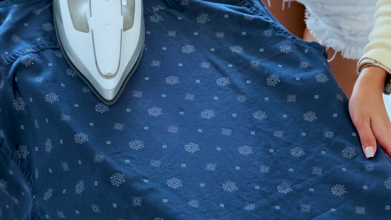 A woman uses a steam iron to press a blue patterned shirt on an ironing board in a brightly lit indoor setting, with smooth camera movement