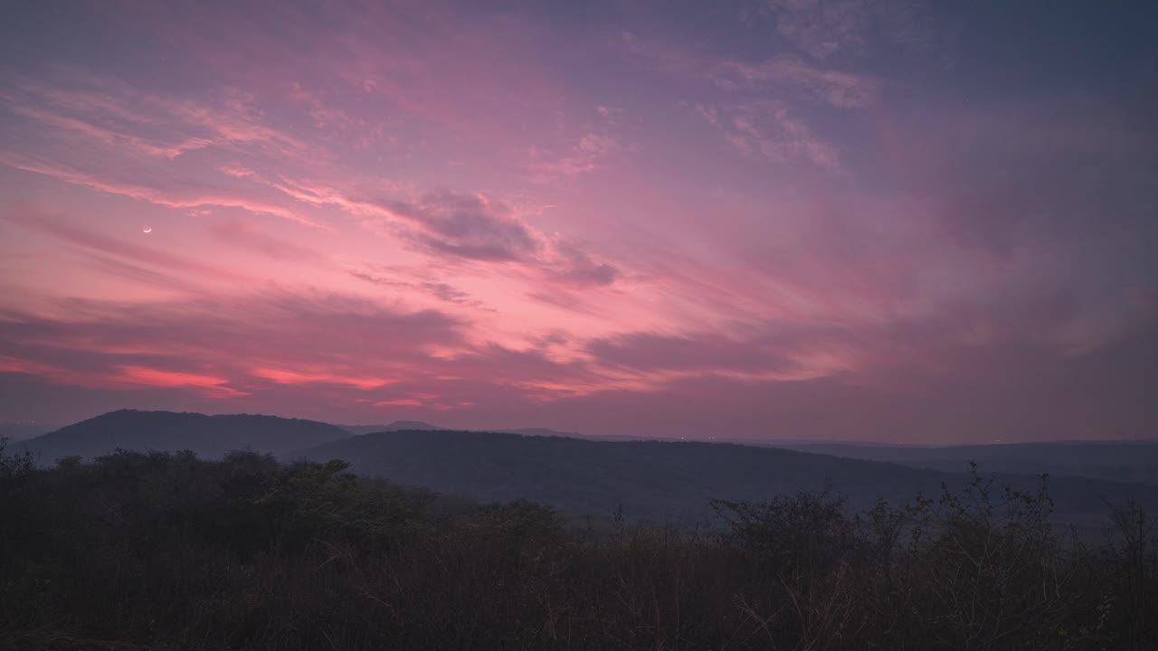 Dag til nat time-lapse af en farverig skumringsaftenhimmel, der smelter sammen med en stjerneklar nat med et bakket landskab og måne i forgrunden