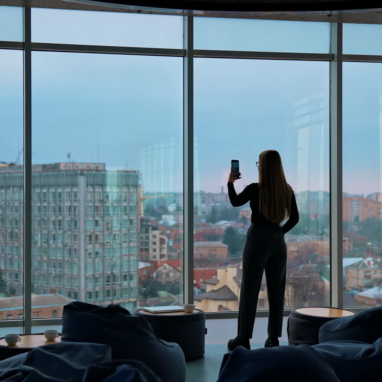 Young woman with a phone near window. Panoramic office view with large windows on city background. Rear view of a girl indoors photographing the evening city landscape