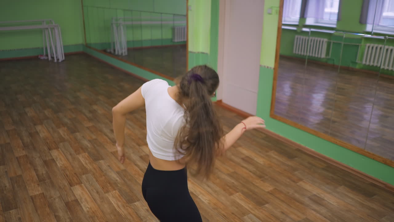 Young lady in white top black leggings and eyeglass dancing in front of mirror, reflection visible on shiny wooden floor, body movement showing rhythm, energy and focus in spacious green hall
