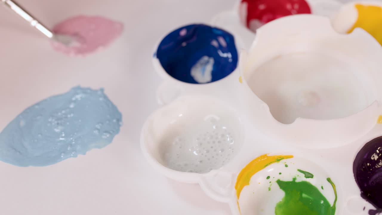 Hand mixes watercolor paints on white palette, creating pastel shades under bright, even lighting