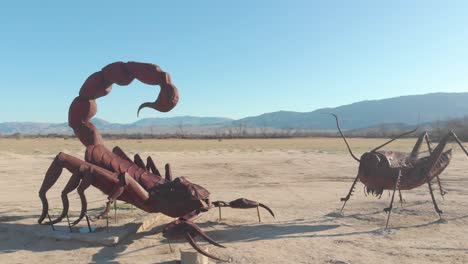 una fotografía de un avión no tripulado de órbita semi-orbital de dos estatuas de un escorpión y un saltamontes mirándose el uno al otro en el desierto con montañas en el fondo