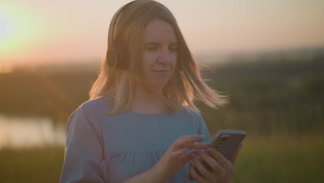 a woman in a blue gown enjoys a sunset, wearing a black headset and using her smartphone, gently nodding to the rhythm