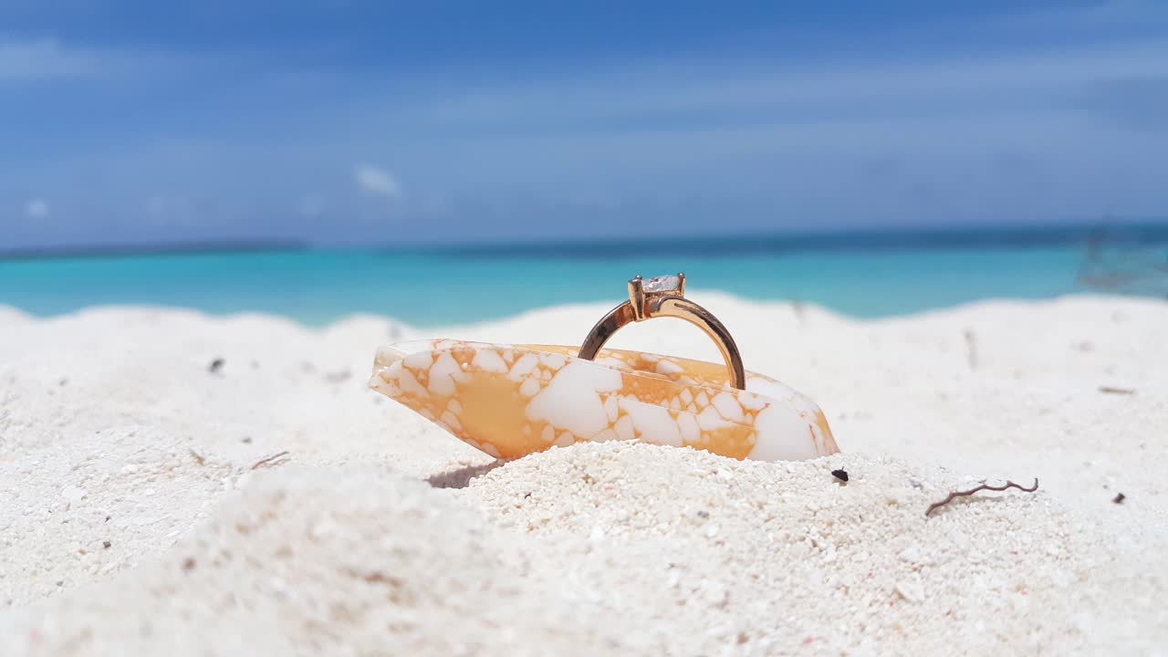Golden bride's ring inserted inside a beautiful seashell over white sand with blur background of azure sea and cloudy sky, concept design