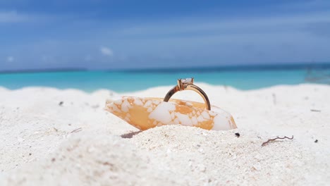 anillo de novia dorado insertado dentro de una hermosa concha sobre arena blanca con fondo borroso de mar azul y cielo nublado, diseño conceptual