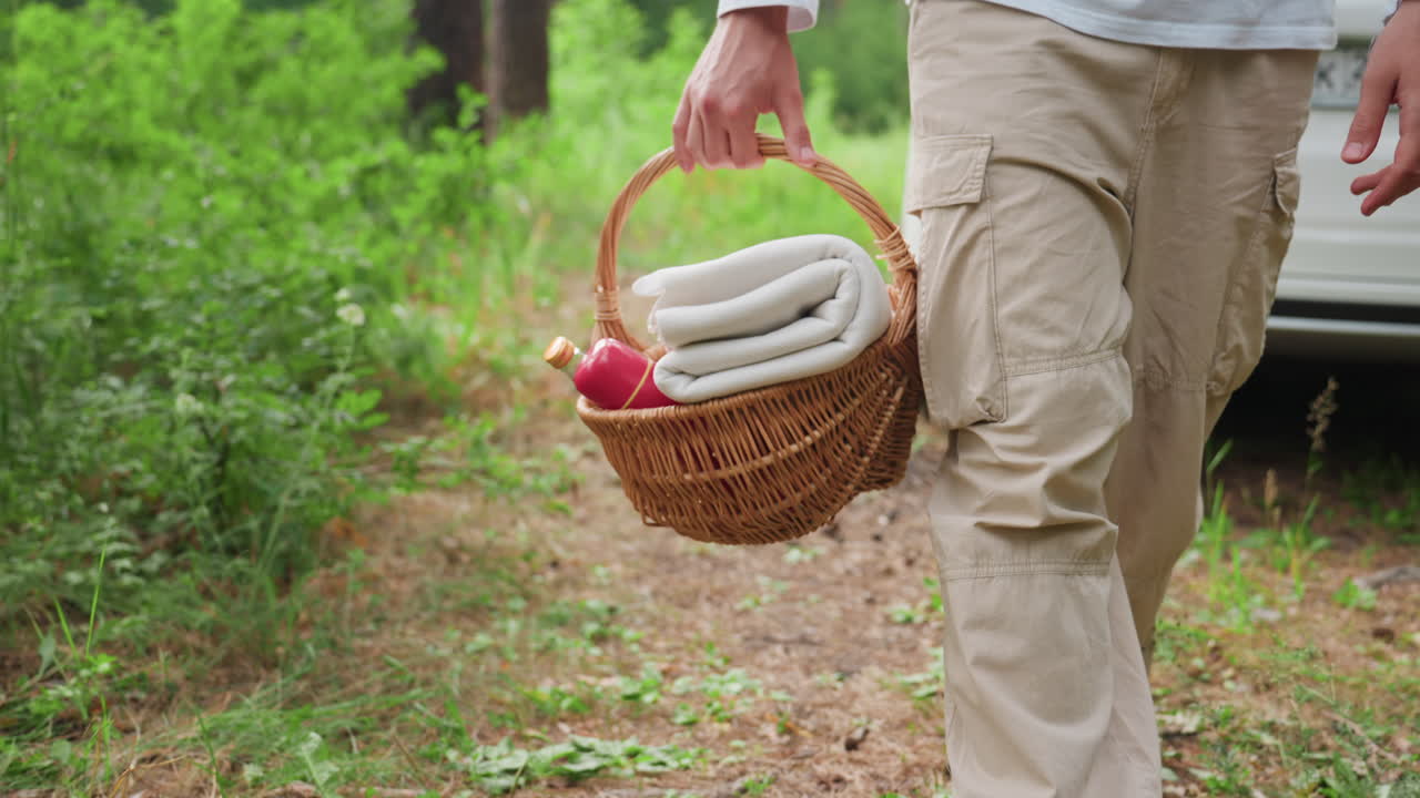 젊은이가 담요와 병이 담긴 고리버들 피크닉 바구니를 들고 흰색 차에서 멀어져 초록색 풀로 둘러싸인 숲길을 따라 걷는 낮은 시점의 모습이며, 평화로운 야외 분위기를 연출해요.