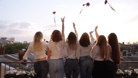 Caucasian-girls-with-beautiful-figures,-in-the-same-casual-clothes-standing-with-their-backs-to-the-camera,-lean-against-the-balcony-railings.-throw-flowers-into-the-air.-Cheerful.-Backside.