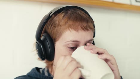 una mujer joven con auriculares beba una bebida caliente mientras ve la computadora portátil en la cama