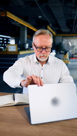 mature caucasian businessman in white shirt and glasses opens his laptop. man starting work in the office. vertical video.