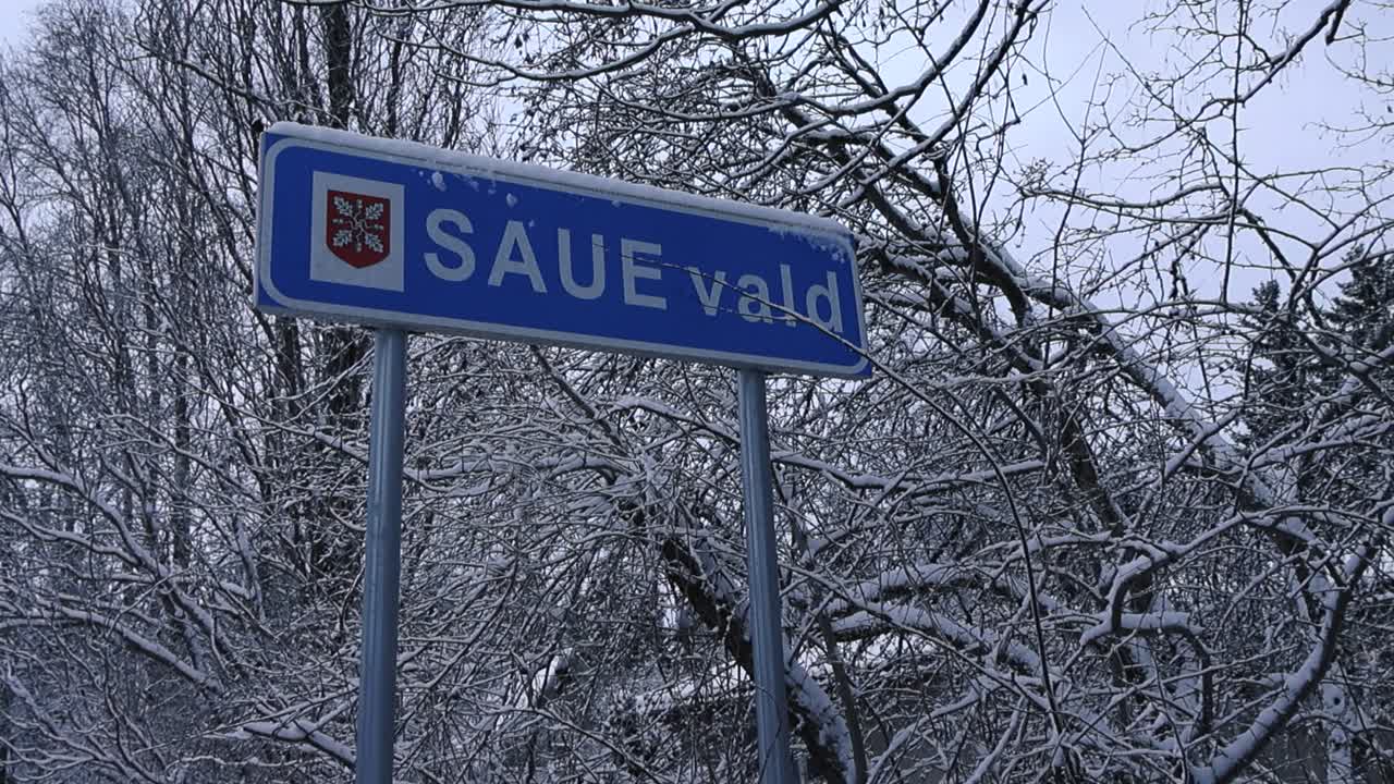Right to left panning footage of a location sign that says SAUE Vald meaning Saue district. The sign is blue and has white edges and is standing between white snow covered trees during winter day time