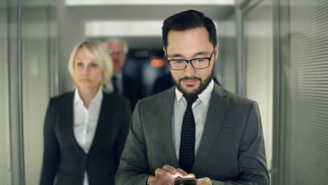 hombre de negocios barbudo camina por un pasillo seguido por su equipo de trabajo mientras escribe en su teléfono inteligente