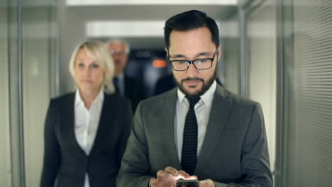 bearded businessman walks through a hallway followed by his work team while typing on his smartphone