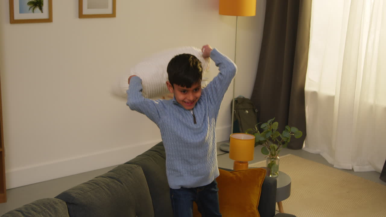 Disruptive Young Boy Behaving Badly At Home Jumping On Sofa And Throwing Cushions Around Lounge