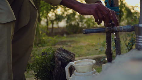 A-man-is-filling-the-ewer-from-an-outdoor-tap-in-the-village,-close-up-view,-a-steel-glass-with-the-tap