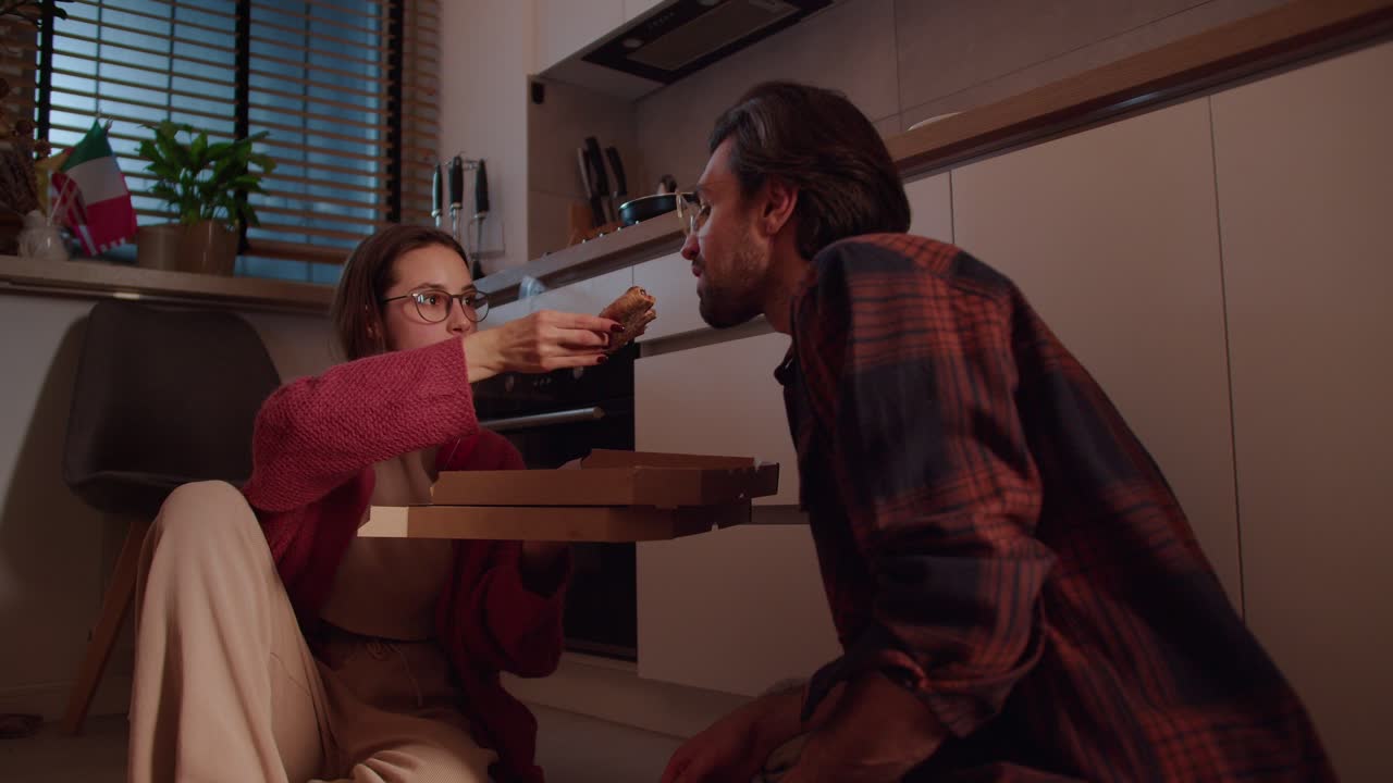 A brunette girl with glasses in a red sweater feeds her brunette boyfriend with stubble pizza from delivery Sitting on the floor in a modern apartment in the kitchen in the evening