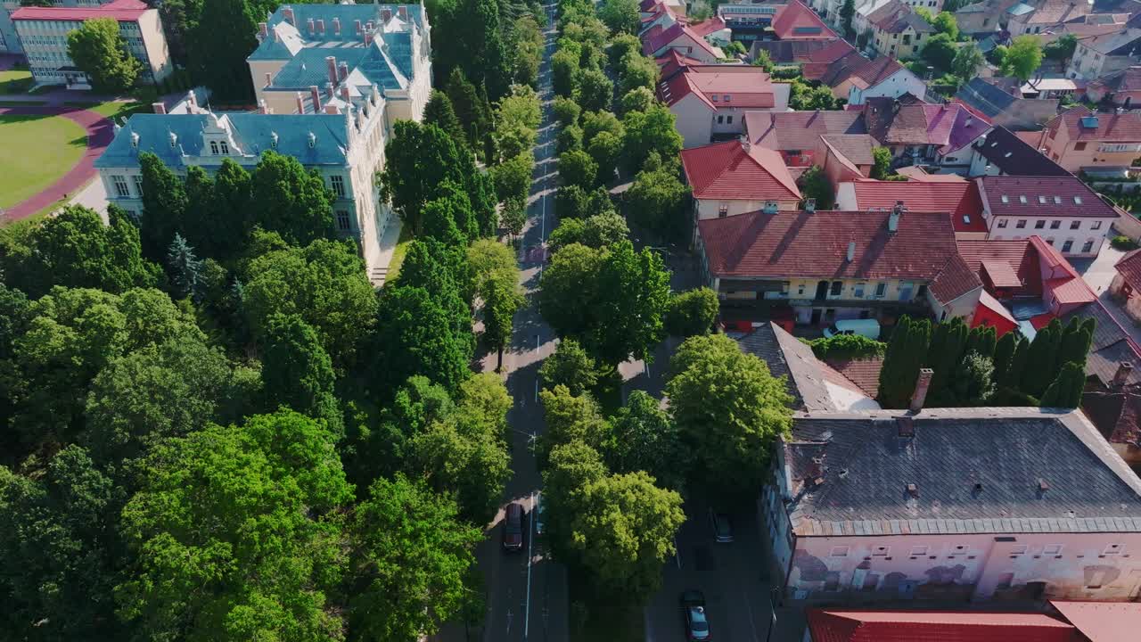 A aerial shot shows a beautiful, green, tree-lined avenue bordered by historic and modern architecture, including a stadium. Light traffic moves through the sunny European town center