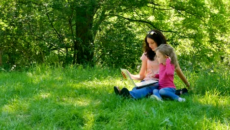 una hija pequeña y bonita con su joven madre están sentadas en la hierba verde en el parque de la ciudad con una computadora portátil blanca, mirando la pantalla y escribiendo algo juntos.