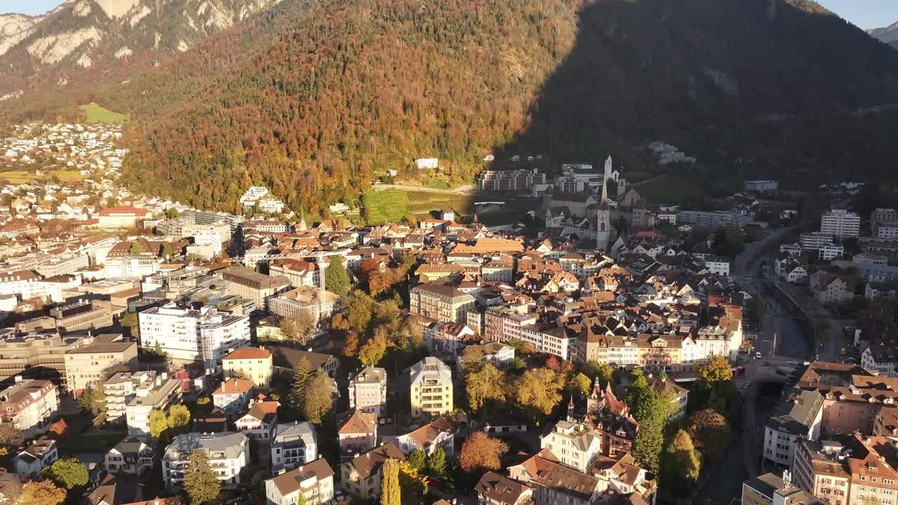 Drone shot moving towards Chur town, bathed in morning sunlight, with majestic mountains in distance, Graubünden, Switzerland.