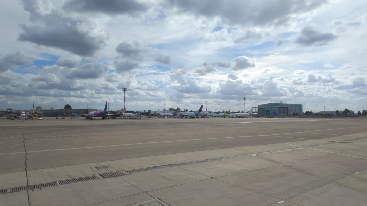 La telecamera scorre verso destra sul piazzale dell'aeroporto, mostrando aerei commerciali parcheggiati e strutture del terminal in condizioni di luce diurna.