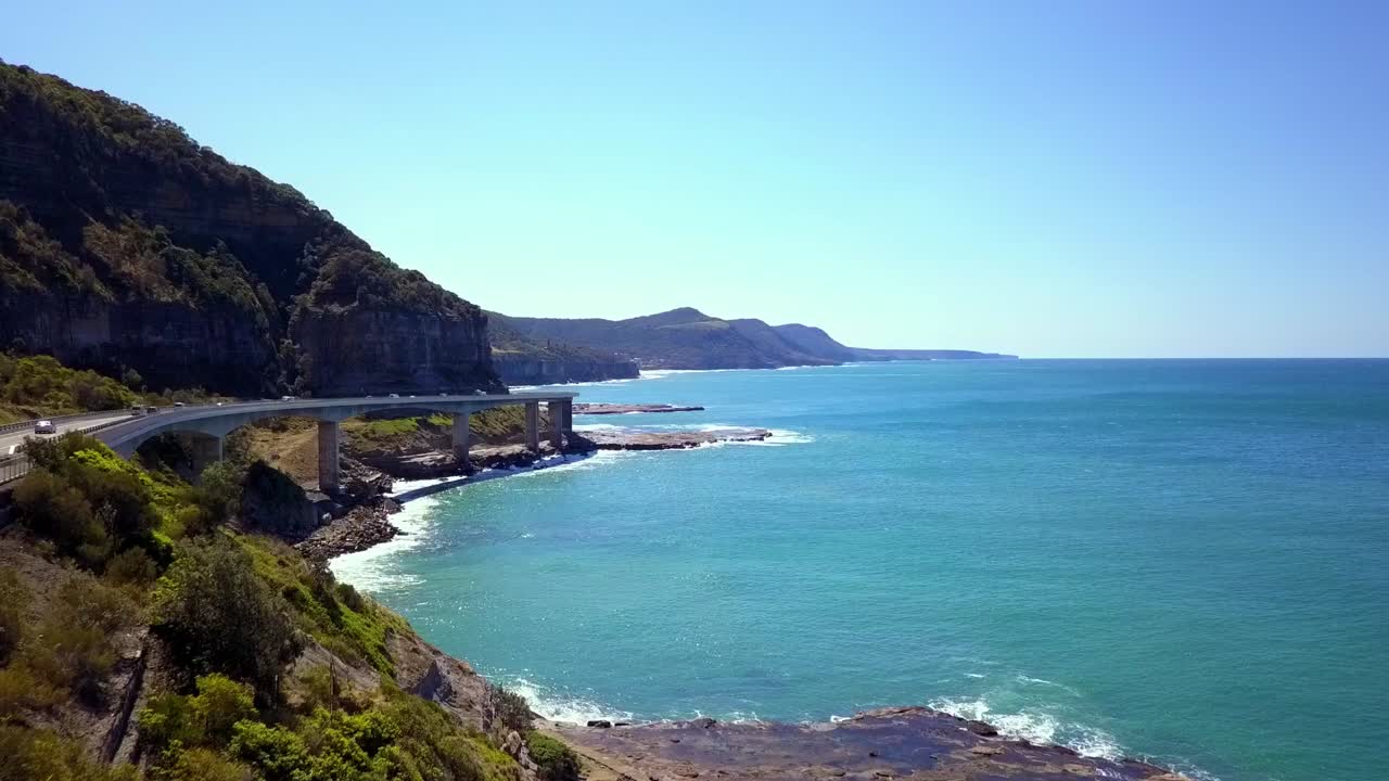 바다 옆에 있는 길고 매력적인 다리의 공중 영상은 호주 시드니의 밝고 화창한 날에 남태평양 바다와 푸른 하늘을 볼 수 있는 그랜드 퍼시픽 드라이브를 강조합니다.