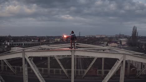 aerial view of young man in hoodie and balaclava standing on the top of the frame construction of an old steel frame bridge with red burning signal flare