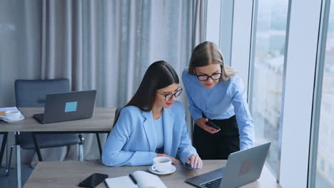 business communication in the modern office. female colleagues looking at laptop communicating and smiling. blurred city panorama in window at backdrop.