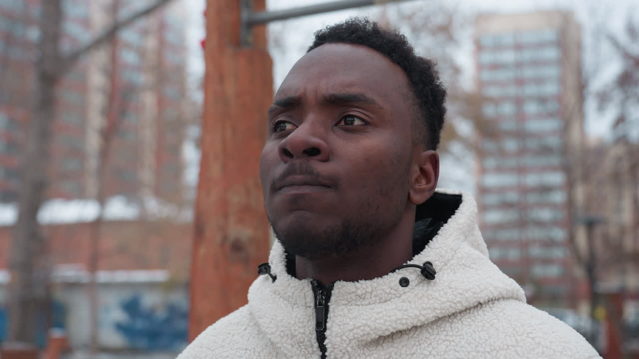 joven decidido que parece centrado mientras está de pie en un área de entrenamiento al aire libre, con una cálida chaqueta de lana, parece profundamente pensativo, el fondo presenta árboles y un entorno urbano