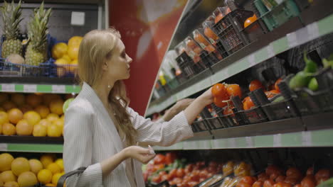 The-girl-housewife-shopping-at-the-supermarket.-Chooses-fresh-tomatoes-on-a-branch-puts-them-in-a-cellophane-bag-ties.-Them-and-puts-them-into-a-cart.