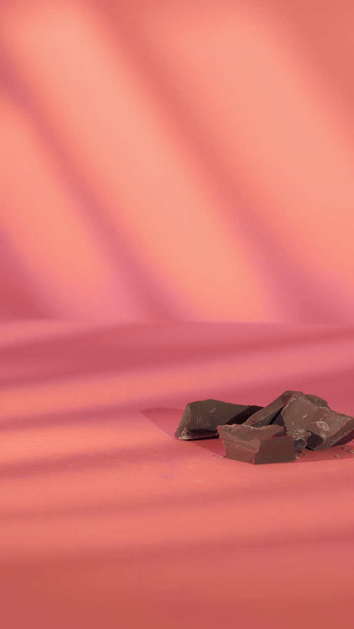 Detailed close-up of a hand reaching to grab a dark chocolate protein bar beside broken chocolate pieces. Soft lighting and pink background create a clean, modern, and appetizing visual.