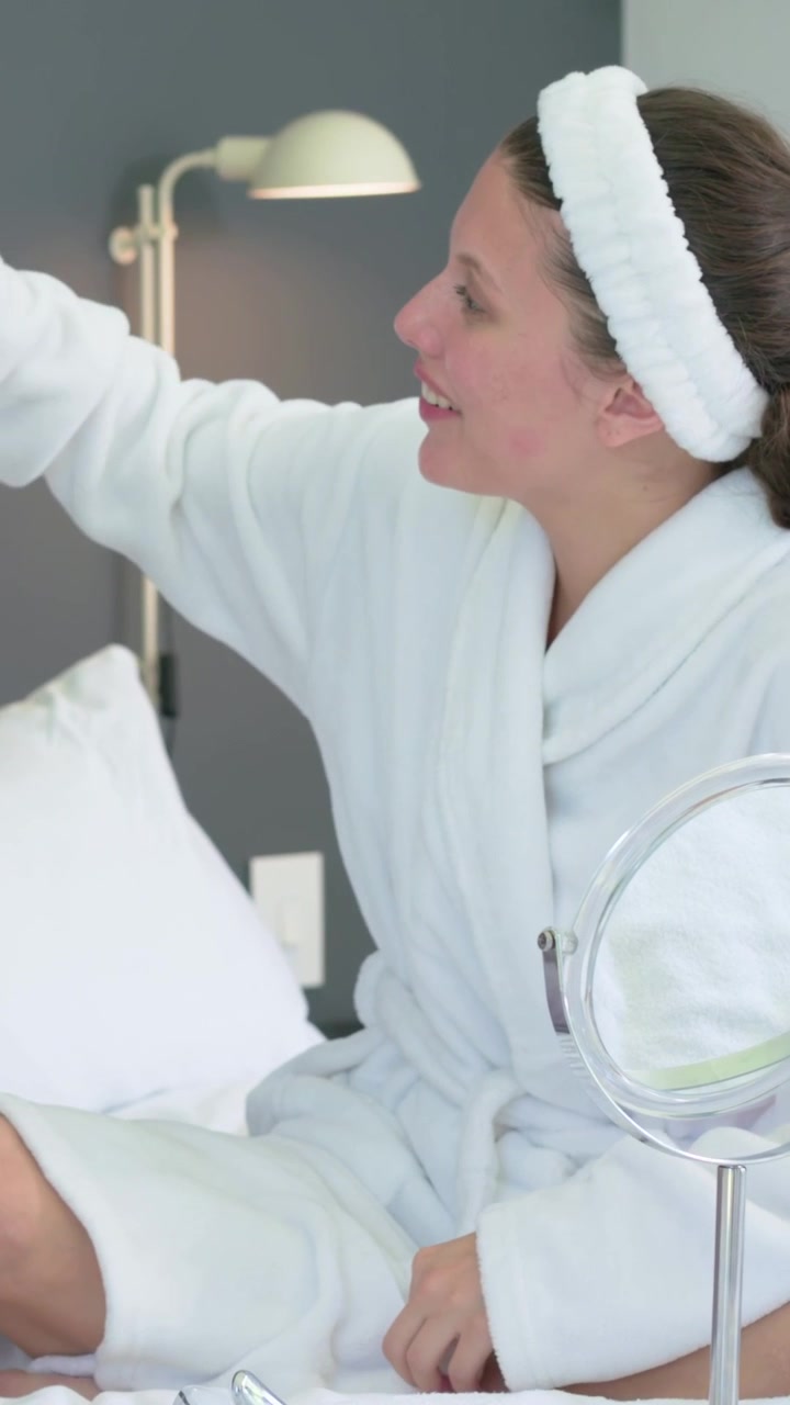 Vertical video: Smoothing hair, woman in robe and headband reaching left on bed with mirror talking