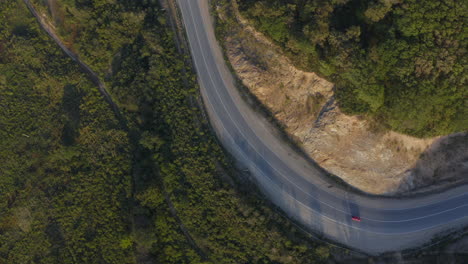 Draufsicht-Auf-Ein-Rotes-Auto,-Das-Bei-Sonnenuntergang-Auf-Einer-Langen-Kurvenreichen-Straße-In-Den-Grünen-Bergen-Mit-Einer-Klippe-Auf-Der-Linken-Seite-Vorbeifährt